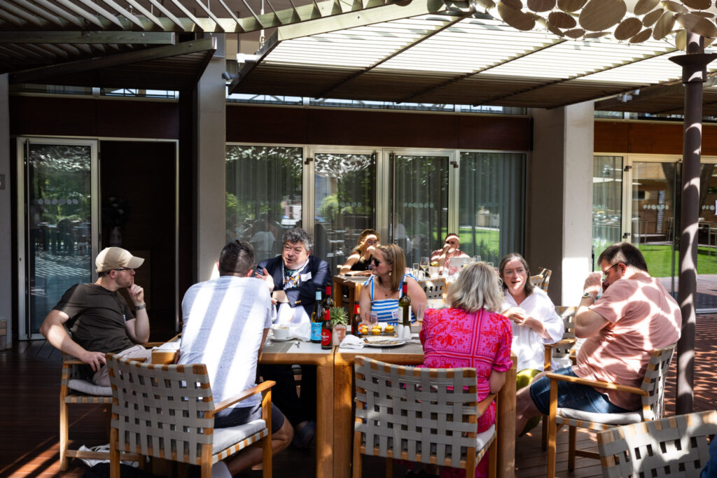 A diverse group of people gathers around a large table outdoors for a meal, enjoying conversation and drinks in a sunlit, casual setting beneath a shaded pergola.
