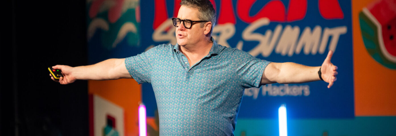 A man with glasses wearing a short-sleeved blue shirt gestures enthusiastically while speaking on stage at the Ibiza Super Summit, with a colorful backdrop featuring the event's name and playful designs.