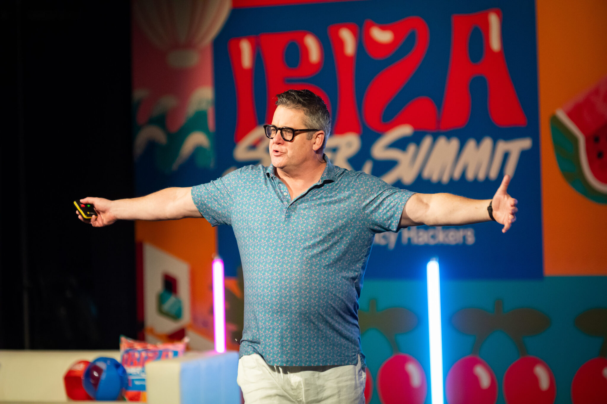 A man with glasses wearing a short-sleeved blue shirt gestures enthusiastically while speaking on stage at the Ibiza Super Summit, with a colorful backdrop featuring the event's name and playful designs.