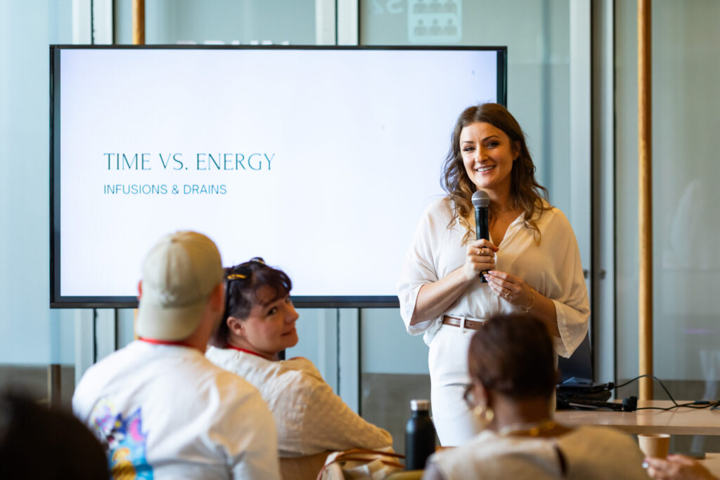 A speaker stands in front of an audience, presenting on the topic "Time vs. Energy: Infusions & Drinks," with a screen behind displaying the title. The audience is engaged, seated, and looking at the speaker.