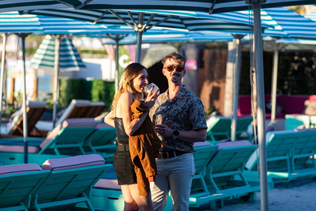 A woman in a black dress and a man in a patterned shirt pose together at a beachside setting, surrounded by deck chairs and umbrellas, enjoying a sunny day.