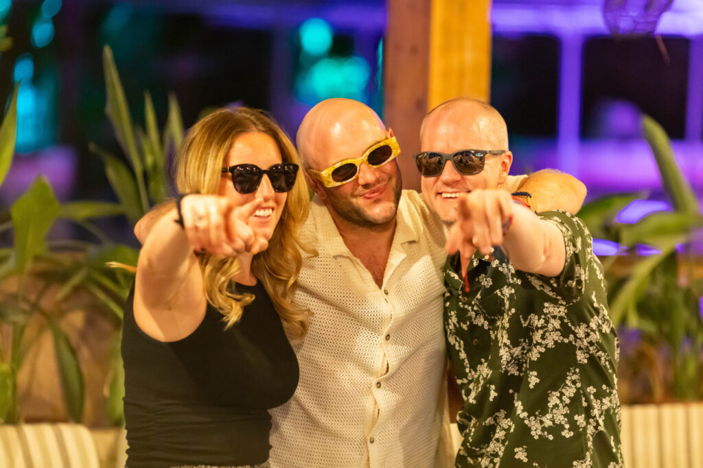 A man and two women stand together, wearing sunglasses and smiling. They are playfully pointing at the camera, surrounded by lush greenery and colorful lighting, suggesting a lively social gathering or celebration.