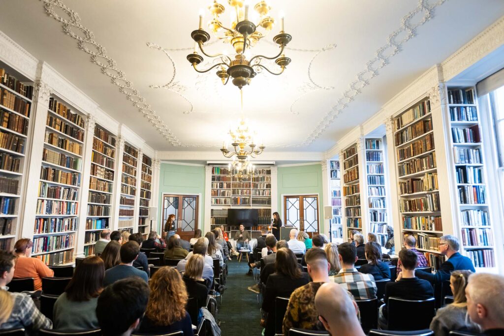 A gathering in a spacious library filled with books, featuring a chandelier and large windows. Attendees are seated, listening intently to speakers at the front of the room, emphasizing a calm and intellectual atmosphere.