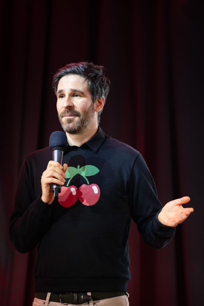 A man with short dark hair stands on stage holding a microphone, wearing a black sweater featuring a cherry design. He appears to be speaking or presenting to an audience, with a red curtain in the background.