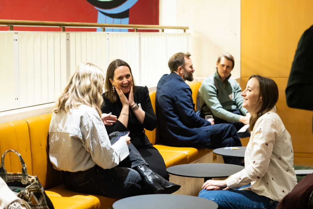 A casual conversation scene featuring four people seated in a lounge area, engaged in discussion. Two women are animatedly talking, while two men observe nearby. The setting has a modern design, with yellow seating and a colorful wall backdrop.