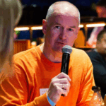 A man with a shaved head, wearing an orange sweater, speaks into a microphone during an event. He appears engaged in conversation, with a blurred background of other attendees.