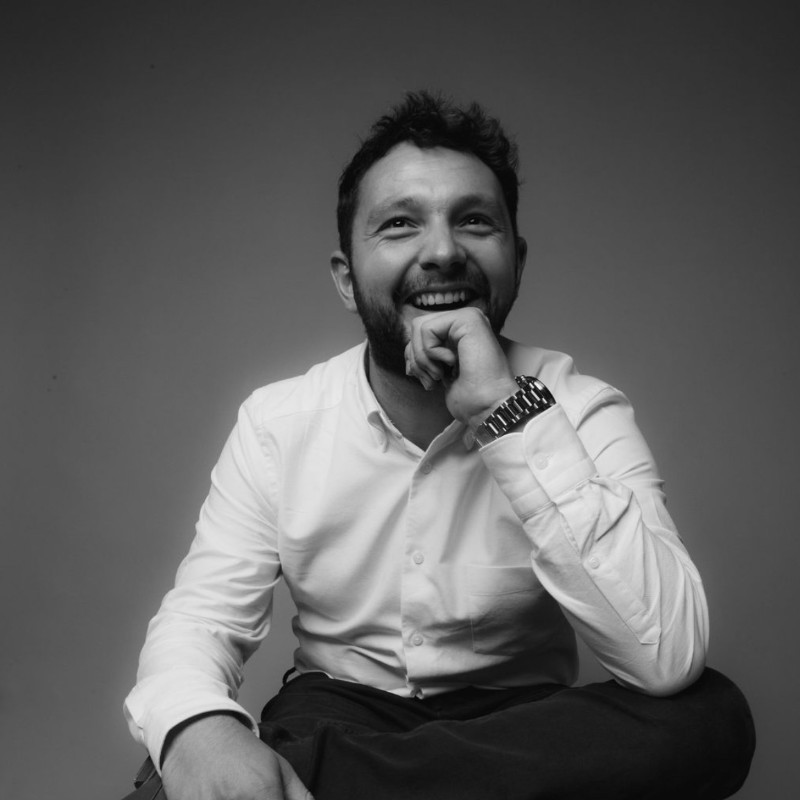 A man with a beard smiles broadly while resting his chin on his hand. He wears a white button-up shirt and sits cross-legged against a neutral background. The image conveys a sense of joy and relaxation.
