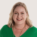 A smiling woman with blonde hair wearing a green sweater is looking directly at the camera. She has a light makeup look and is accessorized with jewelry, exuding a friendly and approachable demeanor.