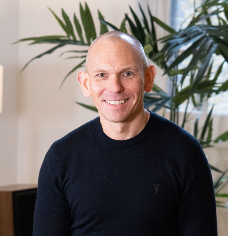 A smiling man with a shaved head is wearing a dark sweater and sitting in a well-lit room with indoor plants in the background.