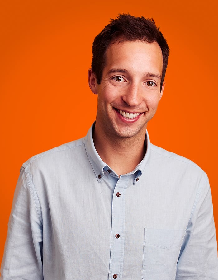 A smiling man with short brown hair is wearing a light blue button-up shirt against a vibrant orange background.