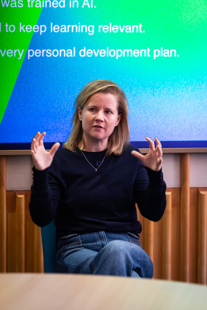 A woman with shoulder-length blonde hair is seated, gesturing with her hands while speaking. She is in front of a colorful presentation screen, discussing topics related to AI and personal development plans in a professional setting.