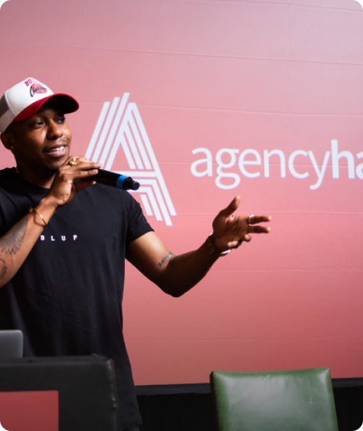 A speaker gestures while presenting at an event, wearing a black t-shirt and a red cap, with a logo for "agency" visible in the background.