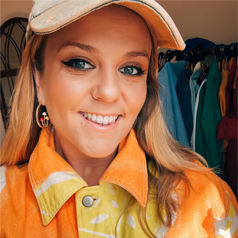 A smiling individual with long blond hair wears an orange jacket with colorful patterns, a white cap, and hoop earrings. The background features hanging clothing items in various colors.