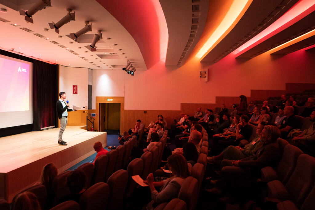 A speaker presents to an audience in a well-lit auditorium, with a large screen displaying visuals. The attendees, seated in rows, are engaged in the presentation. Soft lighting enhances the atmosphere of the event.