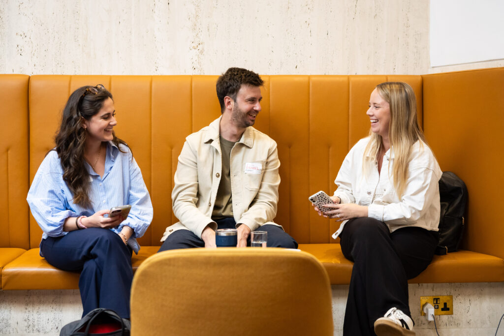 Three young adults sit together on an orange couch, smiling and engaging in conversation. They are casually dressed and holding smartphones, with a glass of water on the table in front of them.