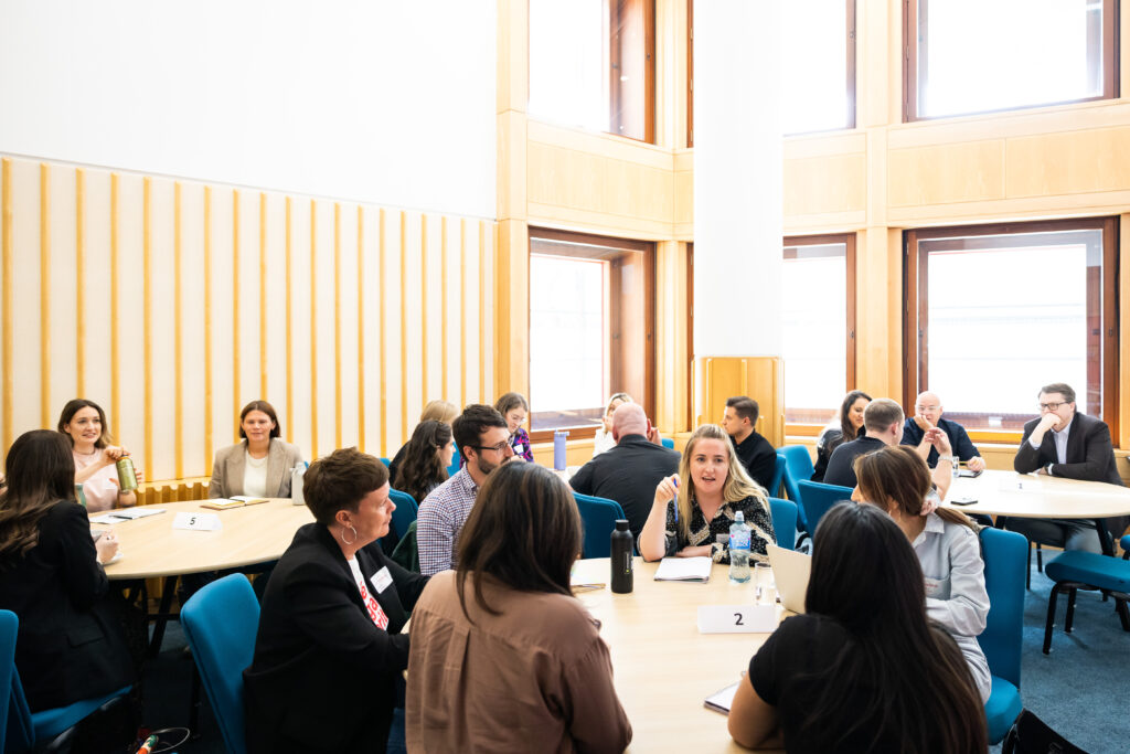 A group of professionals is engaged in discussions around a circular table in a bright, modern meeting room. Various individuals are talking and listening, indicating collaboration and networking during a conference or workshop setting.