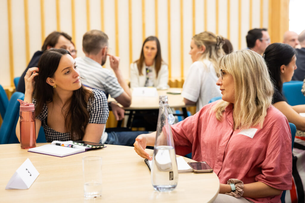 A group of people engage in conversation at a round table in a bright, modern setting. Two women, one in a checked top and the other in a pink blouse, are actively discussing while others are seen in the background.