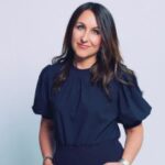 A woman with long, dark hair is wearing a stylish black blouse and poses confidently with a slight smile against a light backdrop.
