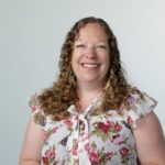 A smiling woman with long, curly hair is wearing a floral blouse. She stands against a light-colored background, radiating warmth and approachability.