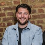 A young man with short dark hair and a beard smiles while seated against a rustic brick wall. He wears a light blue jacket over a black shirt, giving a casual and friendly appearance.