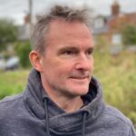 A man with short, light-colored hair is wearing a gray hoodie. He is standing outdoors with a blurred background of greenery and buildings, looking thoughtfully to the side.