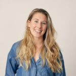 A smiling woman with long, wavy blonde hair is wearing a denim shirt against a light background. She exudes a friendly and approachable demeanor.