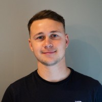 A young man with short hair smiles at the camera, wearing a dark t-shirt. The background is a plain gray wall, creating a simple and neutral setting.