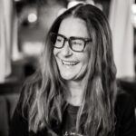 A woman with long hair smiles warmly while wearing glasses. The background is softly blurred, suggesting a lively setting. Her expression conveys happiness and engagement.