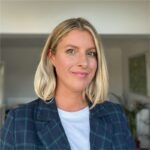 A woman with shoulder-length blonde hair wears a checked blazer and a white shirt. She smiles confidently, with a neutral background that suggests a bright indoor space.