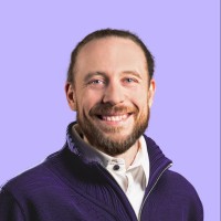 A smiling individual with a beard and long hair, wearing a purple sweater over a white shirt, is portrayed against a light purple background.