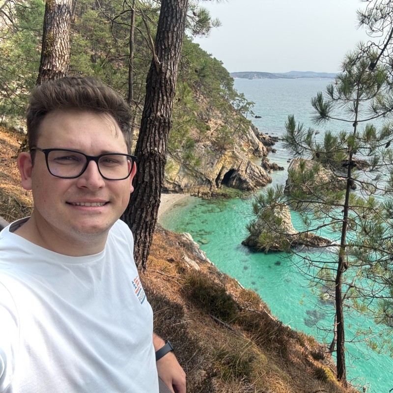 A person wearing glasses and a white shirt taking a selfie on a cliffside with clear turquoise water and rocky coastline in the background, surrounded by pine trees.