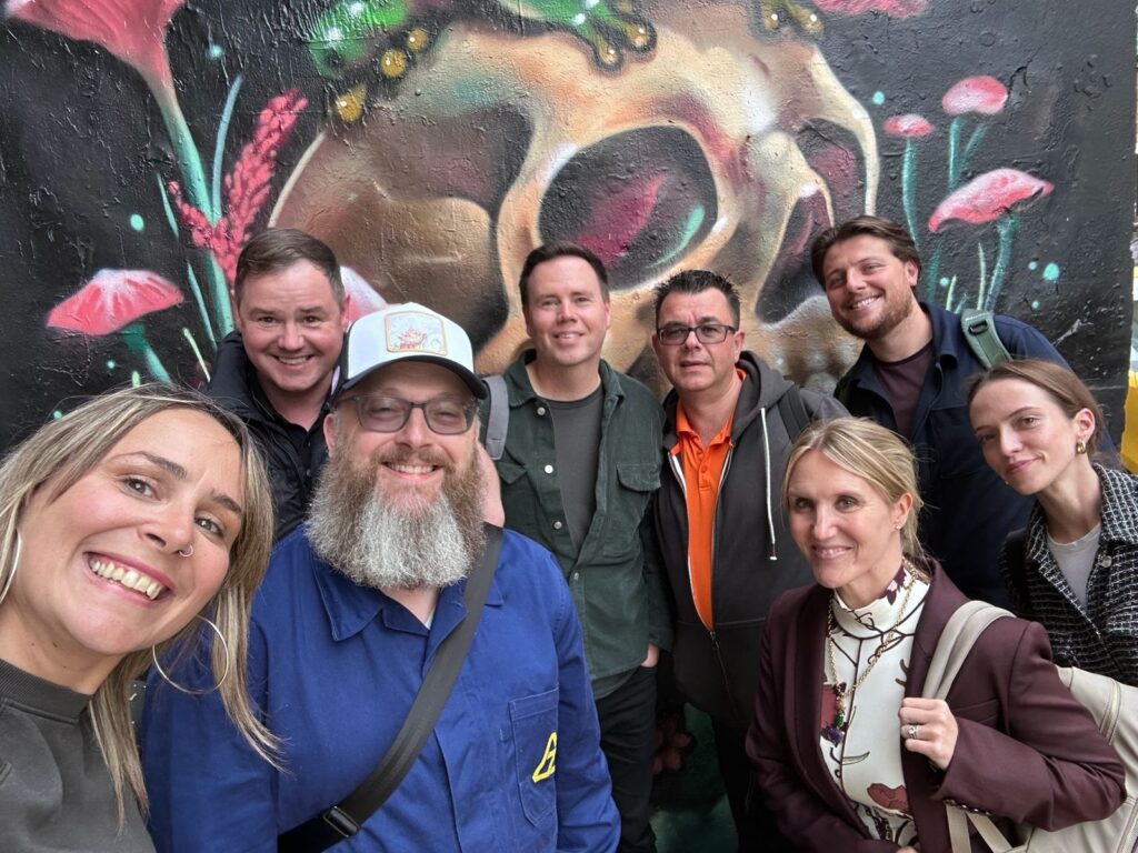 A group of eight people smiles for a selfie in front of a colorful skull mural, surrounded by flowers.
