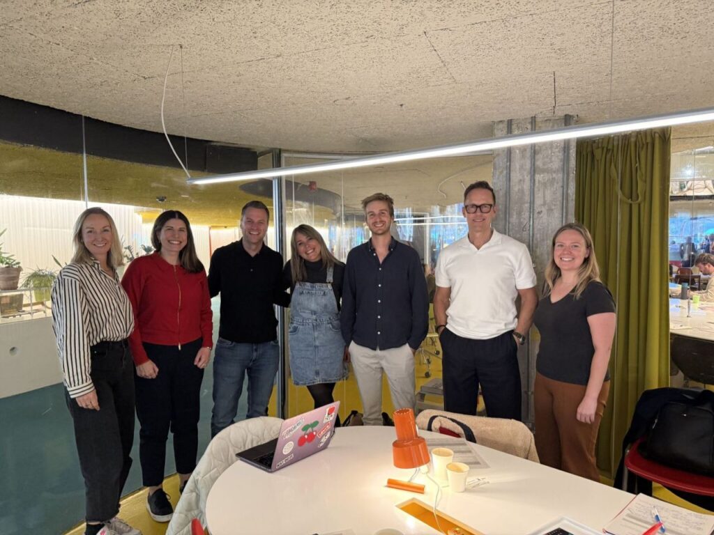 A diverse group of seven people stands together in an office setting, smiling and dressed casually. They are posed in front of a table with various items, including a laptop and a bright orange object. The background features modern design elements.