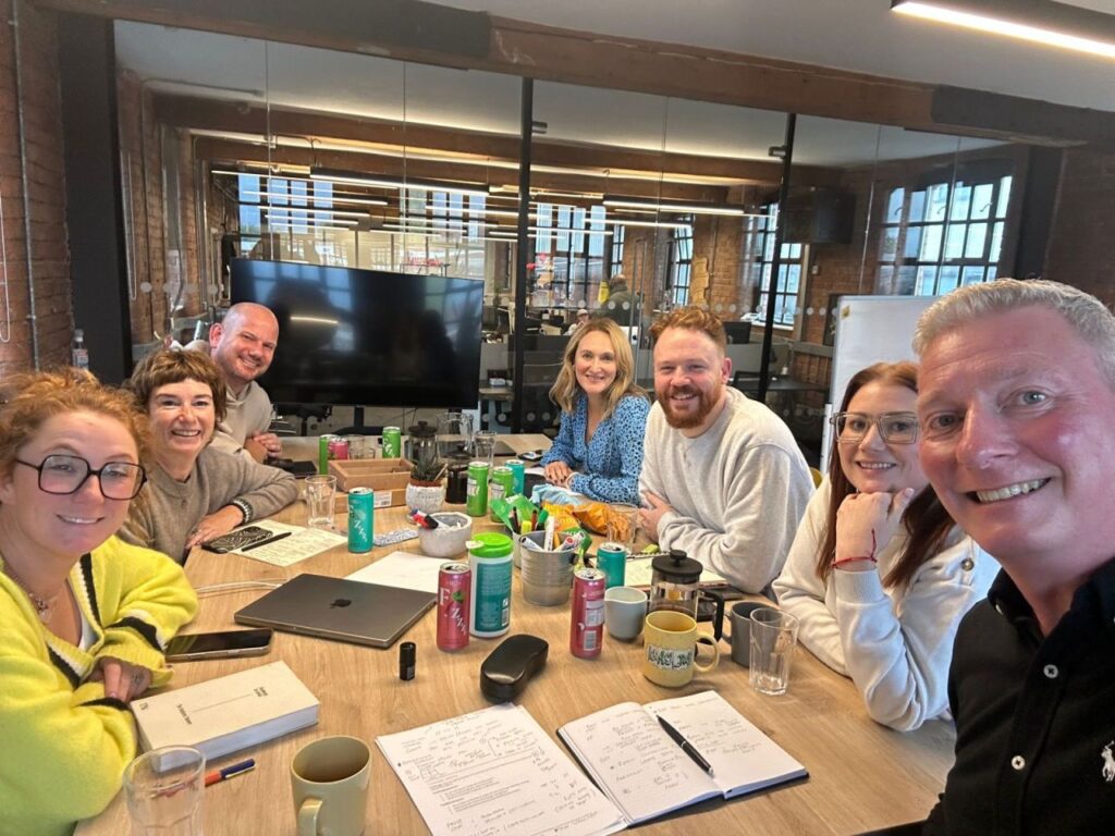 A group of seven individuals is gathered around a conference table with various drinks and notebooks. They are engaged in discussion, with a large screen visible in the background, showcasing a collaborative work environment.