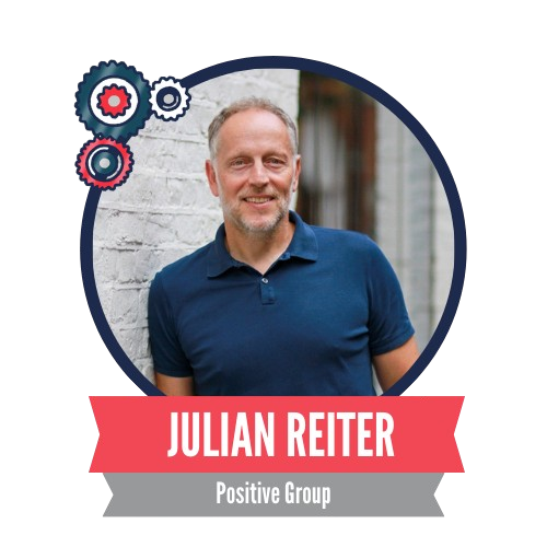 A smiling man in a blue shirt stands in front of a white brick wall. The circular frame features decorative gears, with the text "Julian Reiter, Positive Group" on a red and gray banner below.