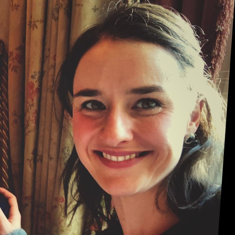 A woman with dark hair smiles warmly at the camera, surrounded by softly patterned curtains. Her friendly expression conveys a sense of approachability and joy.