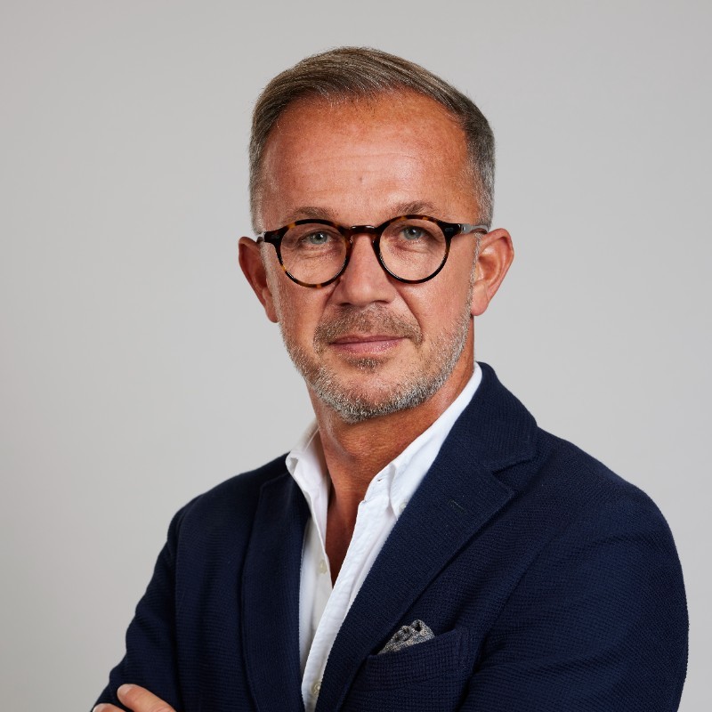A mature man with short gray hair and glasses is posing confidently with his arms crossed. He is wearing a dark blazer over a white shirt, set against a light gray background.