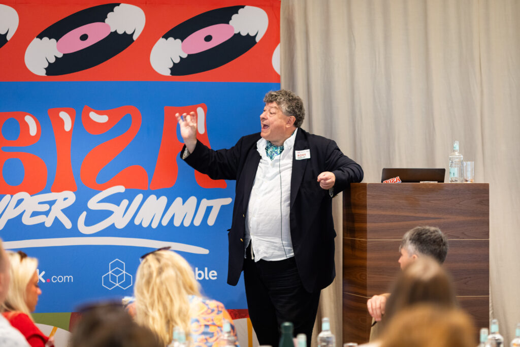 Man presenting enthusiastically at a conference, standing in front of a colorful backdrop with the text "Super Summit." Audience members are seated, listening attentively.