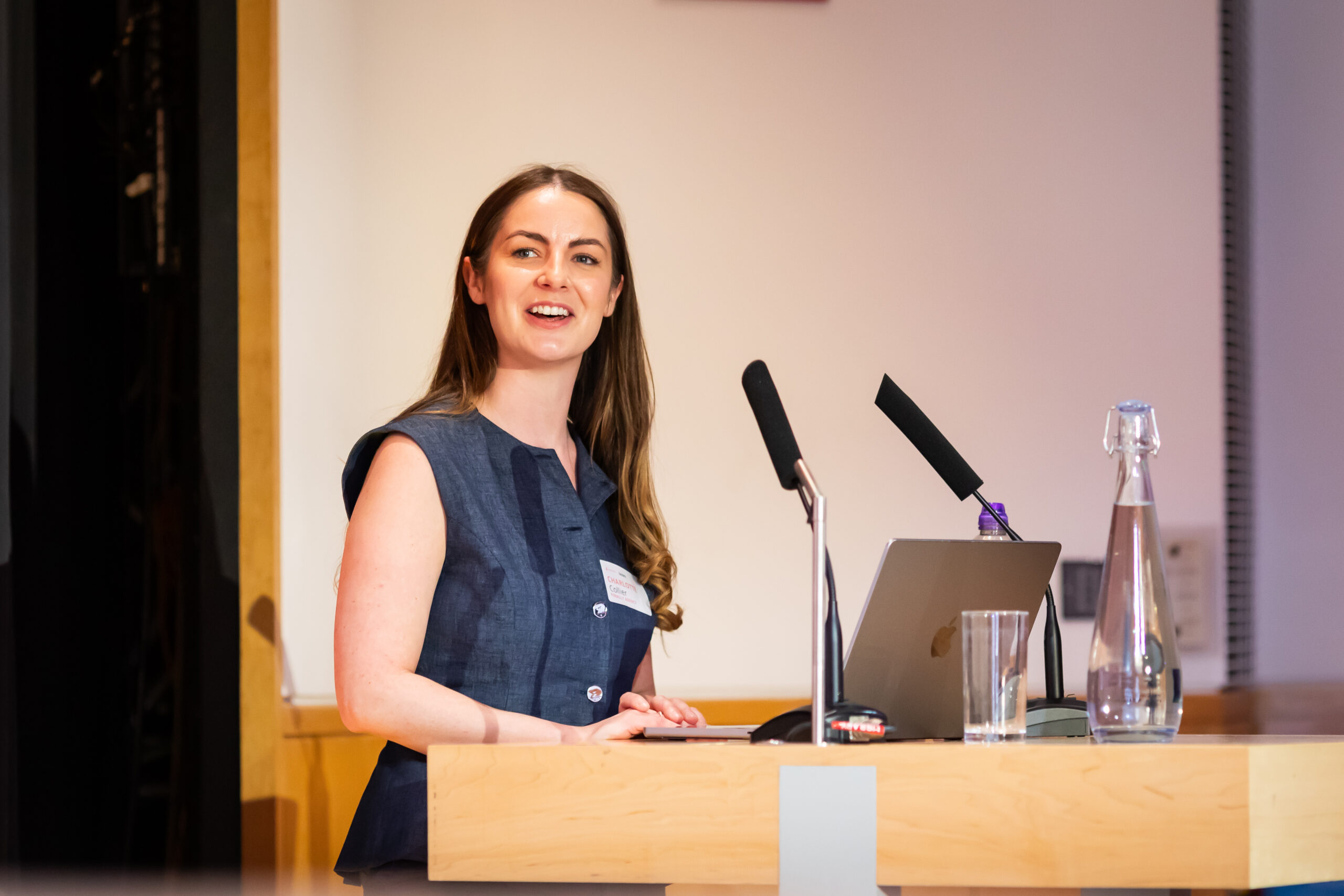 A woman with long brunette hair stands at a podium, smiling while speaking into a microphone. She is wearing a sleeveless blue dress, and there is a laptop and a glass of water on the podium. The background is simple and professional.