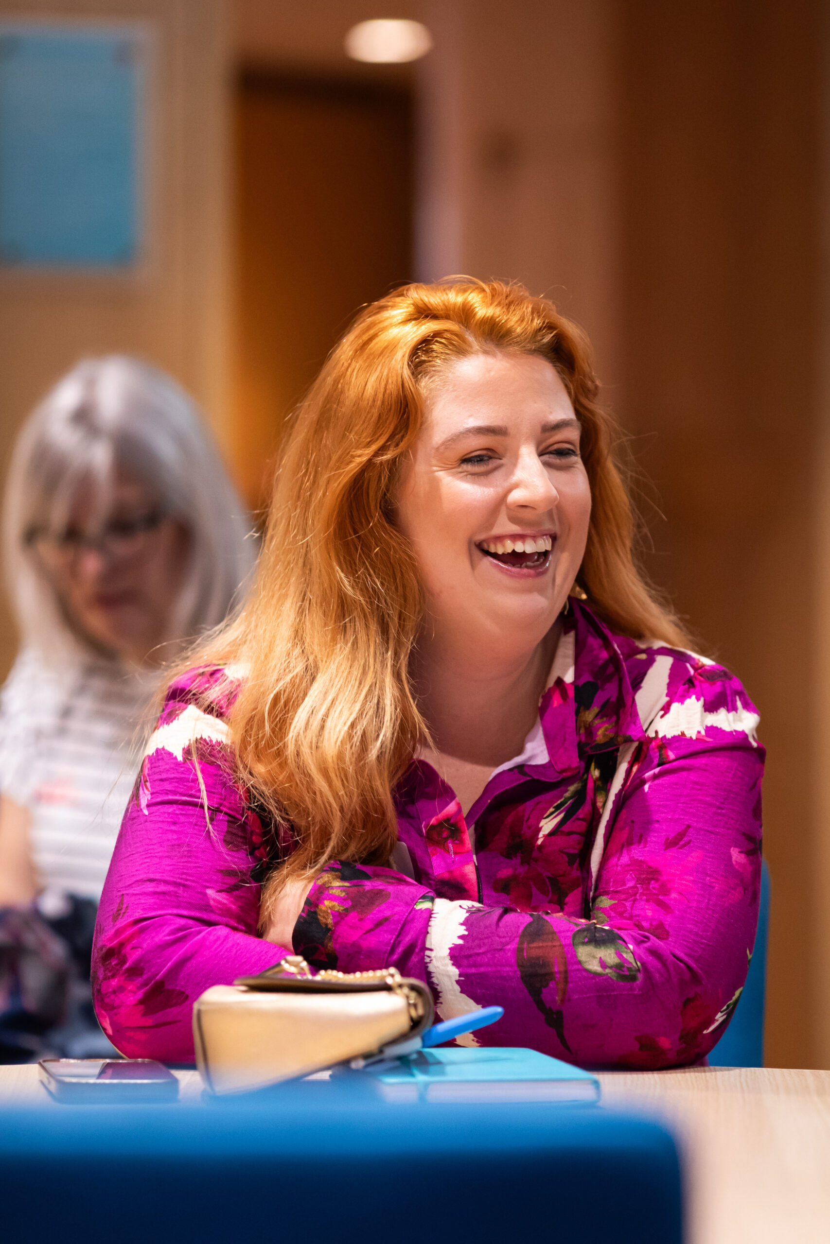 A woman with long red hair and a bright pink floral blouse laughs while seated at a table. In the background, another person is visible, appearing focused on their activity. The setting is casual and relaxed.