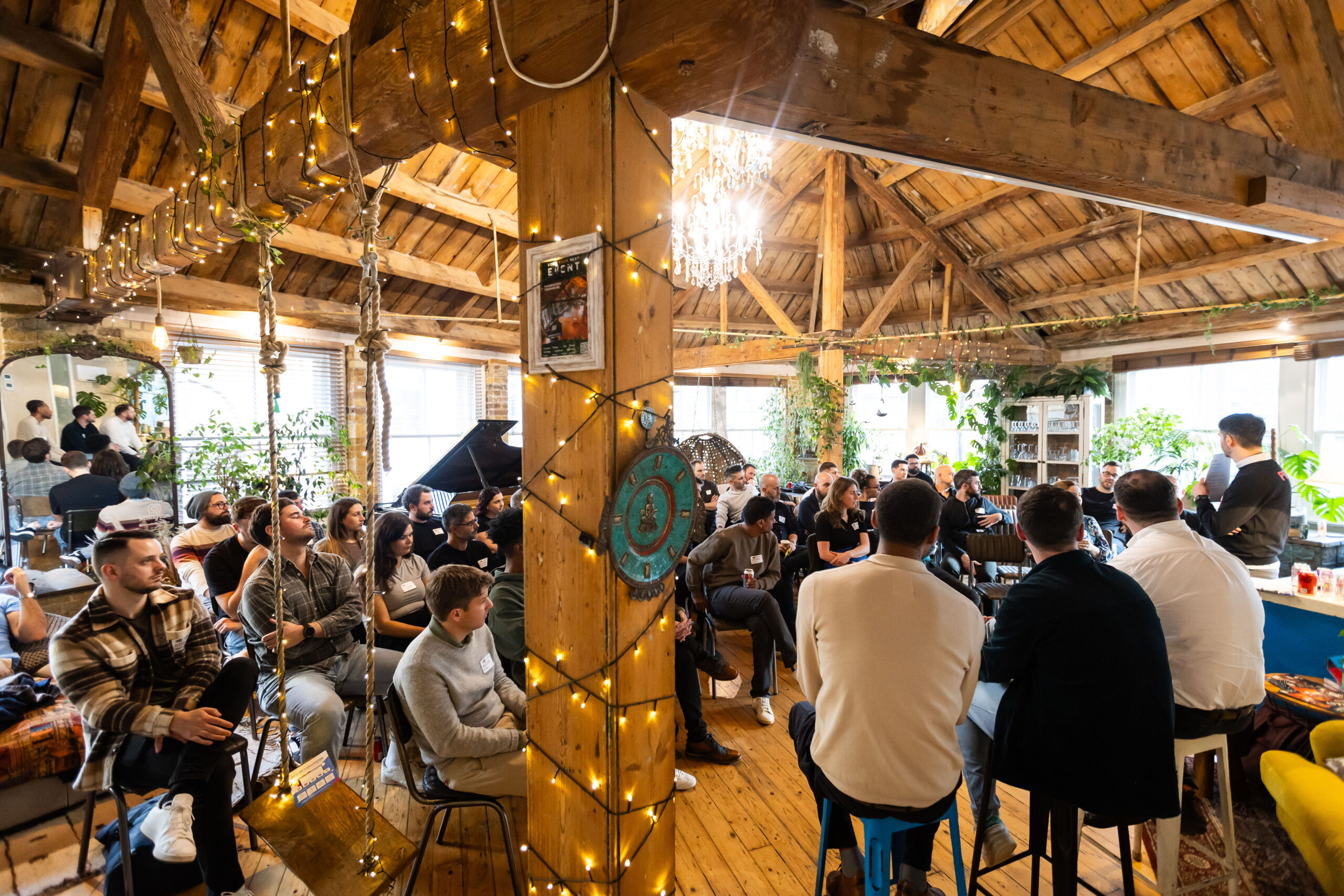 A group of people are seated in a cozy, well-lit space with wooden beams and greenery, engaged in discussion. String lights illuminate the room, creating a warm atmosphere, while participants listen attentively to a speaker at the front.