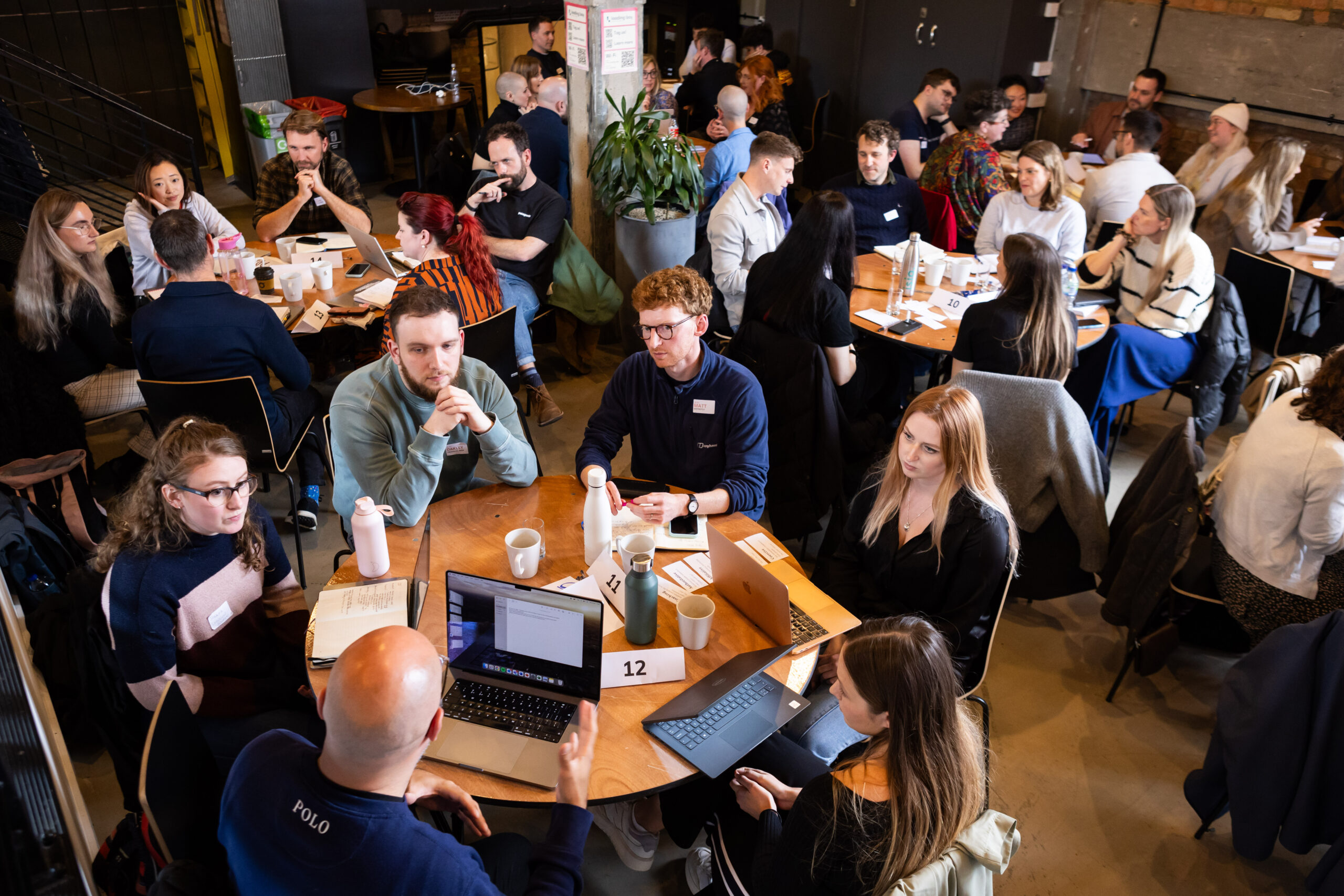 A busy café scene featuring multiple tables with groups of people engaged in conversation. Laptops and notebooks are visible, suggesting collaboration or discussion. The atmosphere is lively, with plants and a modern interior design in the background.