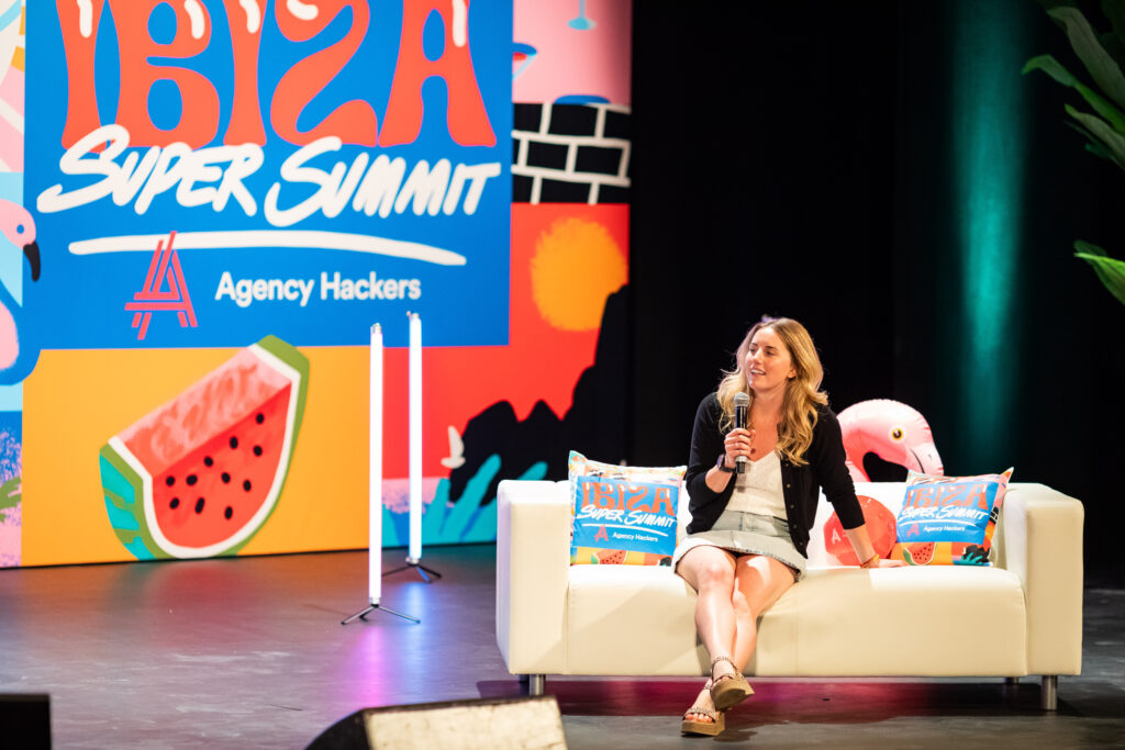 A woman speaks on stage at the Ibiza Super Summit, with a colorful backdrop featuring tropical designs and the event logo. She is seated on a sofa adorned with decorative pillows, engaging the audience with her presentation.