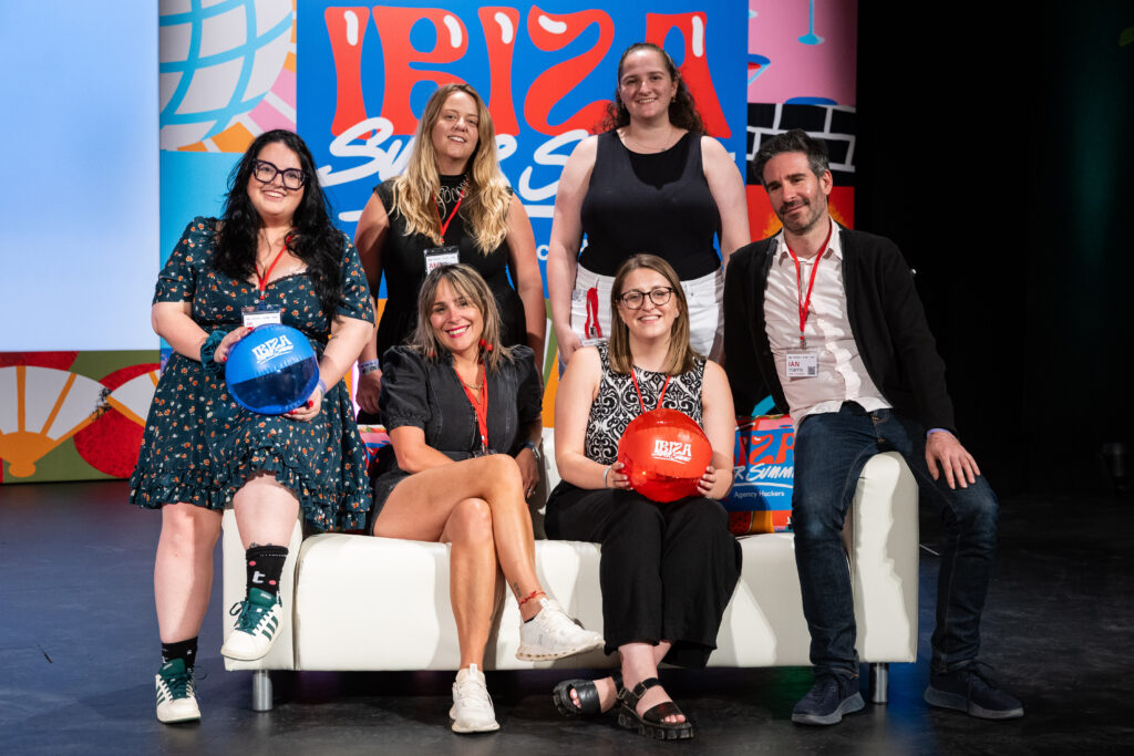 A group of six individuals poses on a couch, smiling and holding colorful balls. They are on stage with a vibrant backdrop featuring designs related to Ibiza Super. The scene conveys a sense of camaraderie and excitement.