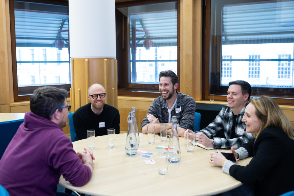 A group of five people sits around a round table in a brightly lit room, engaging in conversation. They have drinks on the table and appear to be enjoying a collaborative discussion.