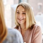 A woman with long blonde hair smiles warmly while engaged in conversation, sitting in a bright, modern setting. Her expression conveys friendliness and openness.
