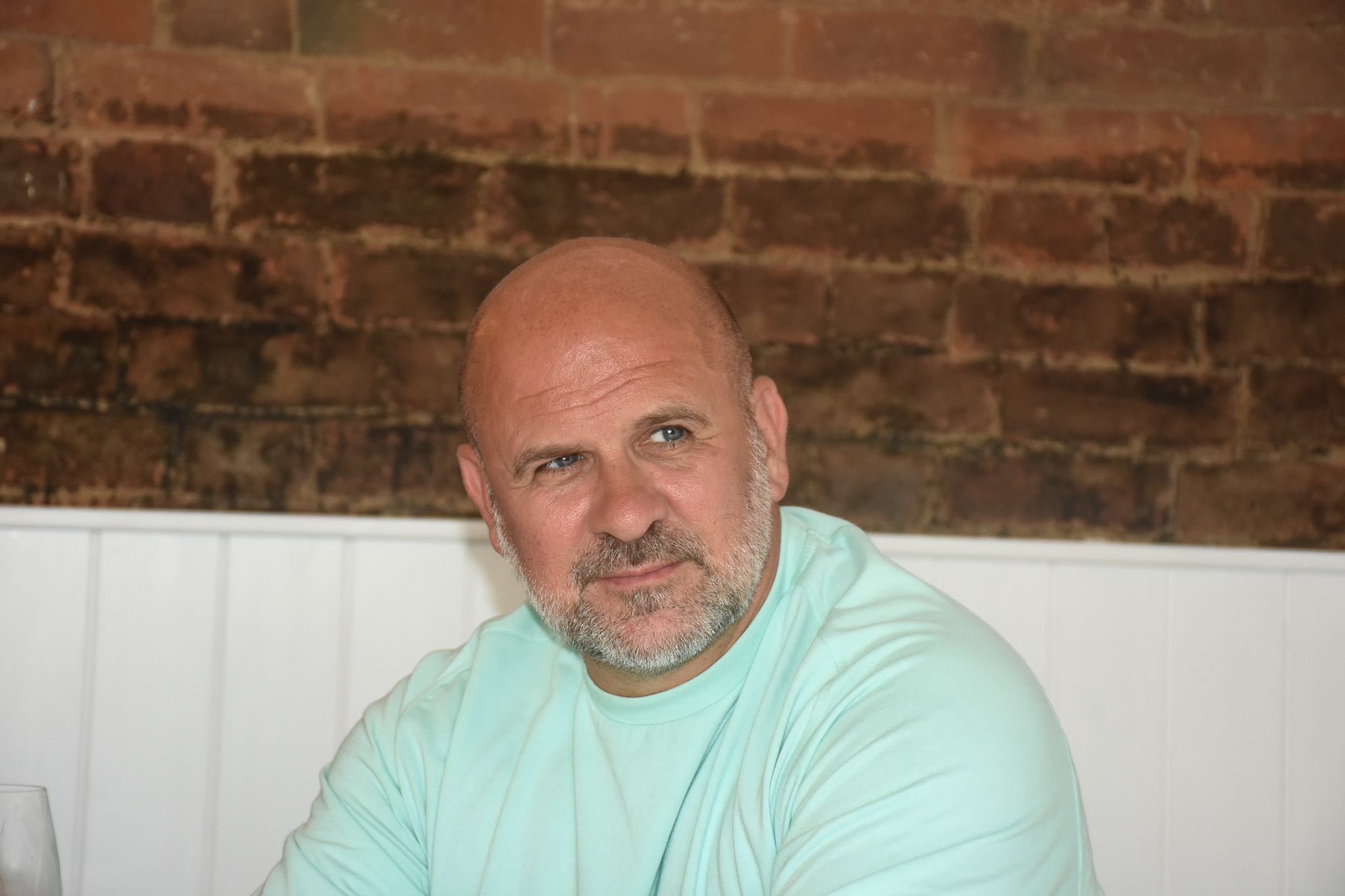 A middle-aged man with a bald head and a beard is sitting in a casual setting, wearing a light turquoise shirt. He looks contemplative, with a brick wall in the background.