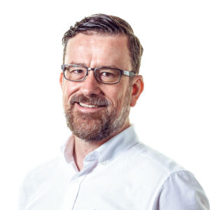 A smiling man with a beard and glasses, wearing a white collared shirt. The background is plain white, emphasizing his friendly expression.