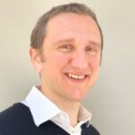 A man with light brown hair and a friendly smile is pictured. He is wearing a navy blue sweater over a collared white shirt, standing against a light gray background.