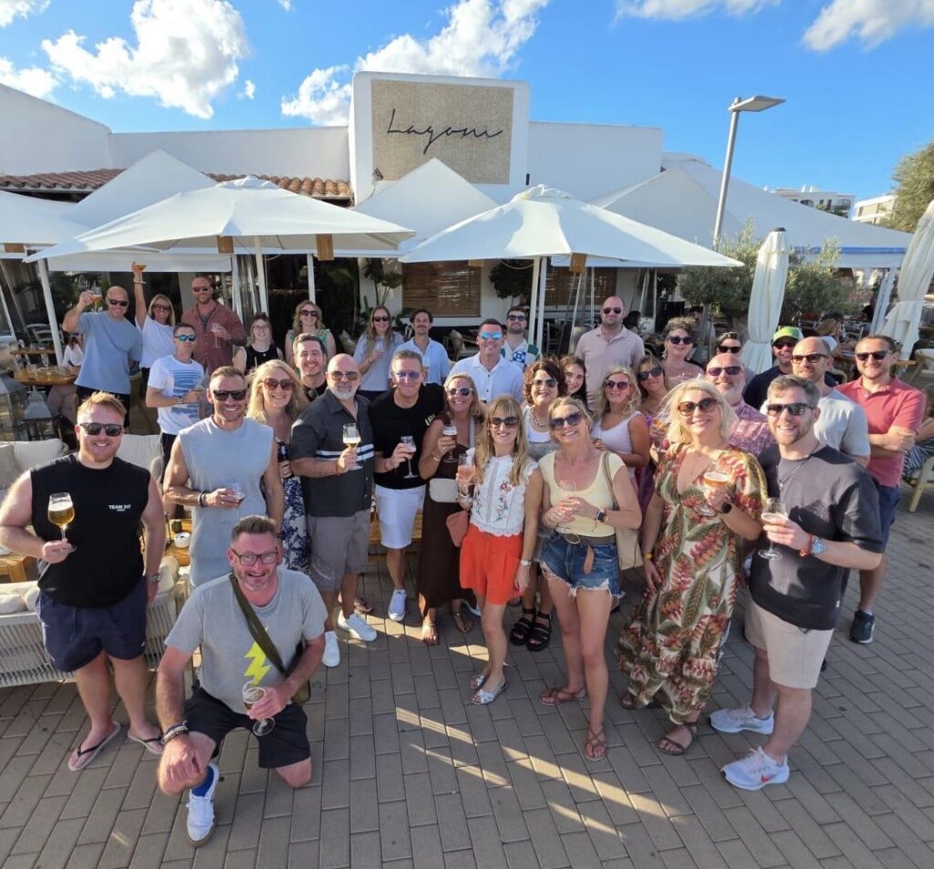 A group of people enjoying a casual outdoor gathering with drinks near a restaurant, under sunny skies and umbrellas.